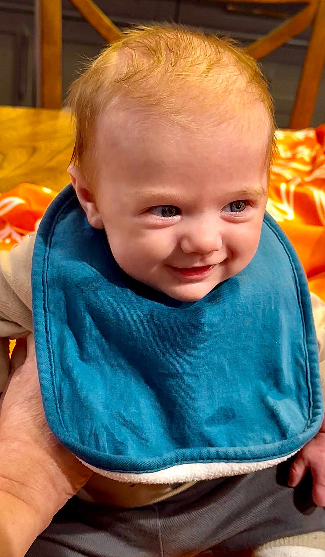 Benjamin joined the competition — help win amazing prizes! baby, child, smile, blue_bib, hand, indoor, wooden_chair, orange_blanket, cute, portrait, clothing, person, infant, happy, face, head, toddler, sitting, young_child, warm_lighting