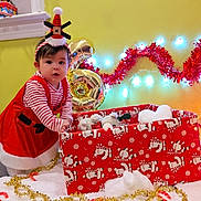 Tita Aylani joined the competition — help win amazing prizes! toddler, child, holiday, christmas, festive, red_dress, striped_shirt, plush_toy, gift_box, candy_cane, christmas_tree, lights, decorations, hat, indoors, yellow_wall, table, candle, celebration, cute