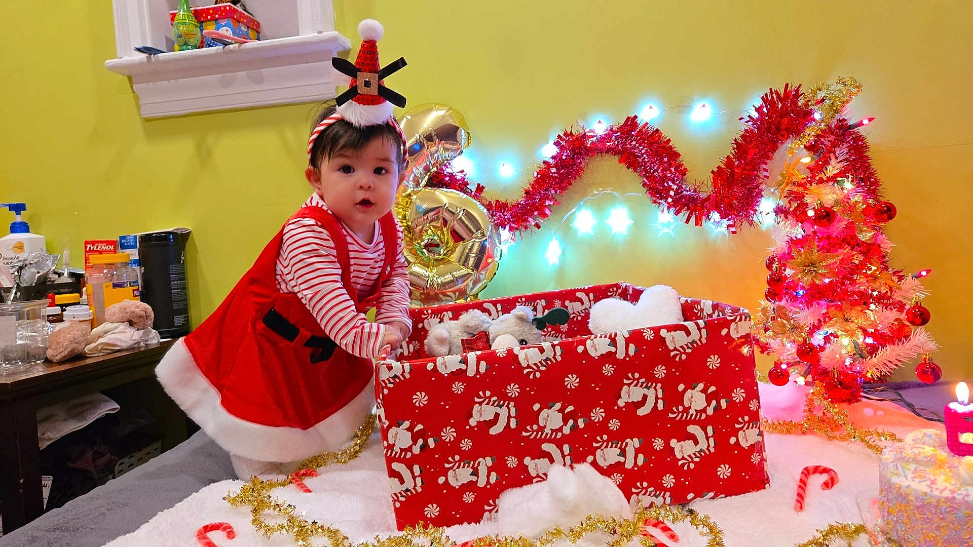 Tita Aylani joined the competition — help win amazing prizes! toddler, child, holiday, christmas, festive, red_dress, striped_shirt, plush_toy, gift_box, candy_cane, christmas_tree, lights, decorations, hat, indoors, yellow_wall, table, candle, celebration, cute