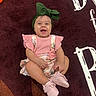 baby, infant, child, smiling, happy, portrait, sitting, green_bow, headband, pink_outfit, floral_clothing, pink_booties, rug, indoor, fashion, cute, face, full_body, toy, patterned_background