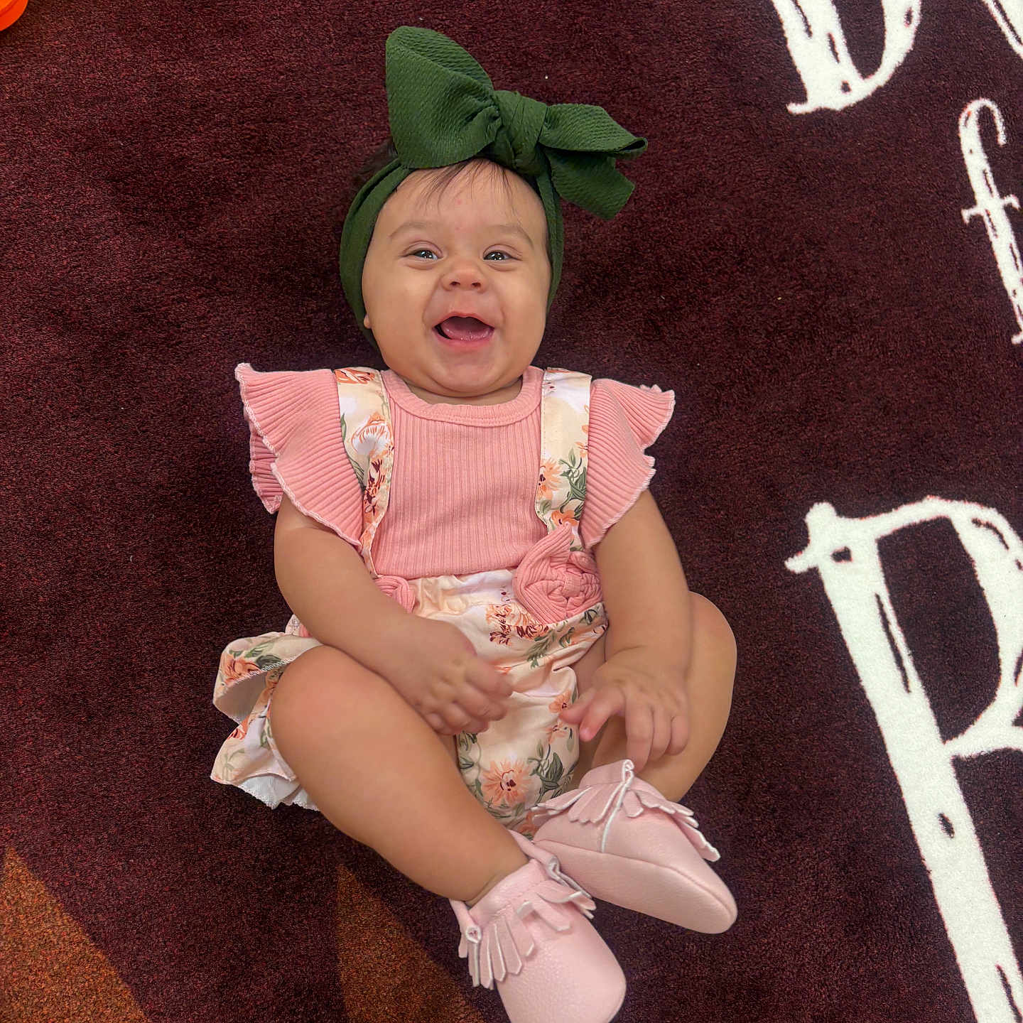 Jeanlle is registered to the contest to win money with this photo: baby, child, cute, face, fashion, floral_clothing, full_body, green_bow, happy, headband, indoor, infant, patterned_background, pink_booties, pink_outfit, portrait, rug, sitting, smiling, toy