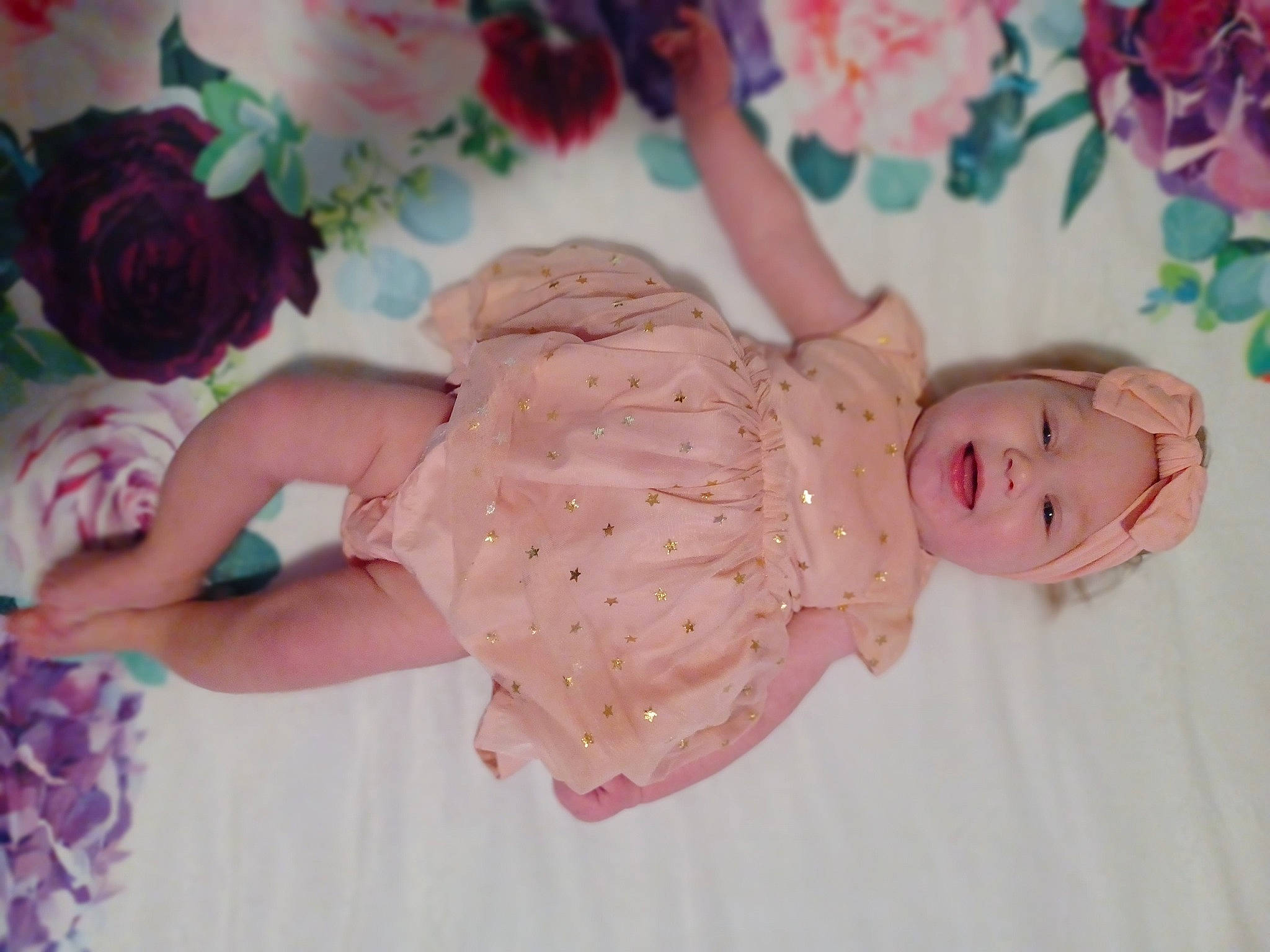 Delaney is registered to the contest to win money with this photo: arm, baby, baby_toddler_clothing, child, dress, flower, hair, happy, headpiece, headwear, human_body, leg, pattern, peach, person, petal, pink, plant, rose, textile