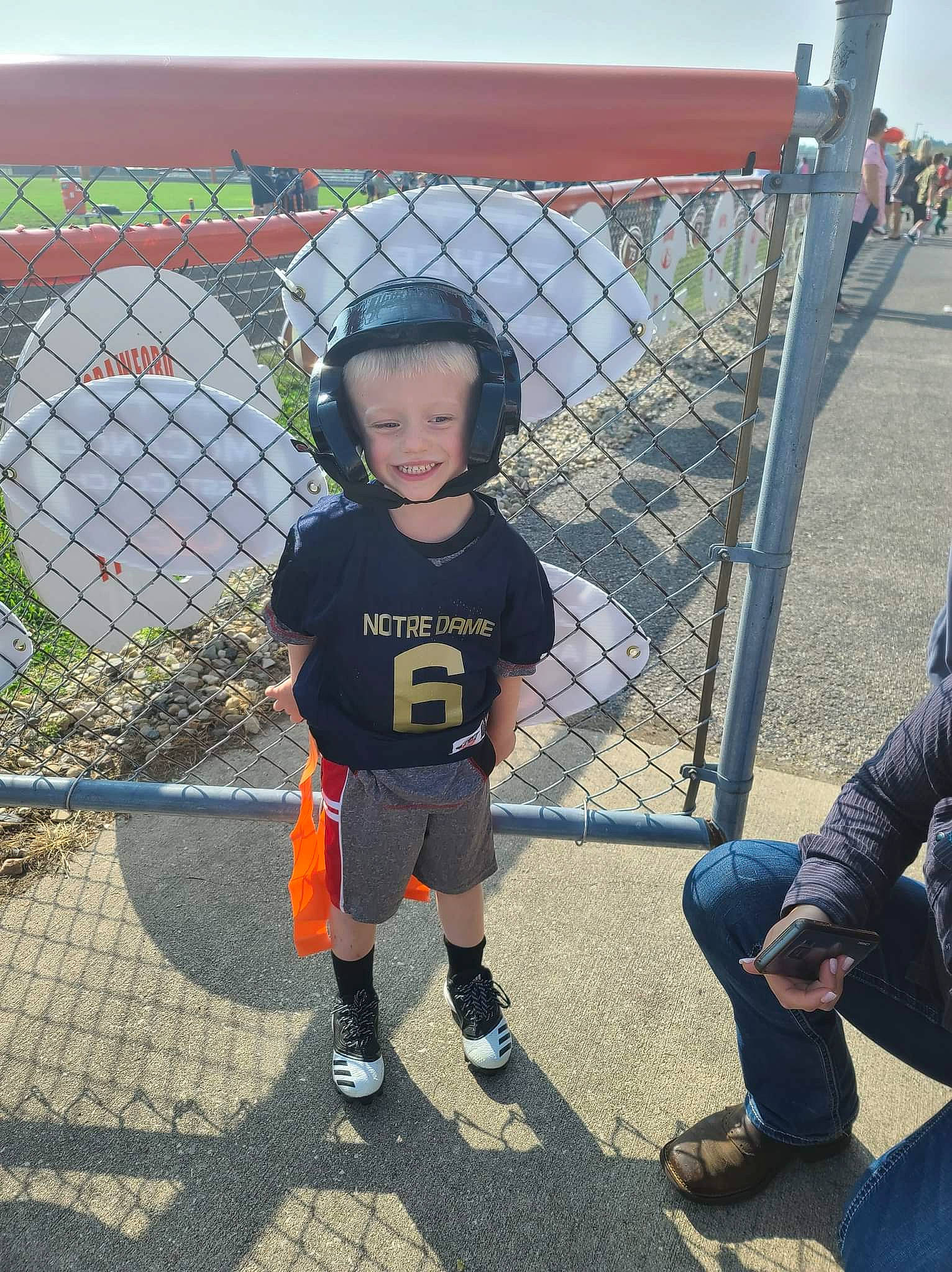 Zane is registered to the contest to win money with this photo: fence, footwear, fun, grass, hat, joy, leg, leisure, mesh, person, personal_protective_equipment, player, recreation, sandal, shoe, shorts, smile, sneakers, sports_equipment, toddler