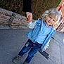child, toddler, blond_hair, curly_hair, denim_jacket, blue_shirt, jeans, smile, holding_hand, adult_hand, boots, sidewalk, brick_wall, street, houses, urban, outdoor, sunny, portrait, casual_clothing