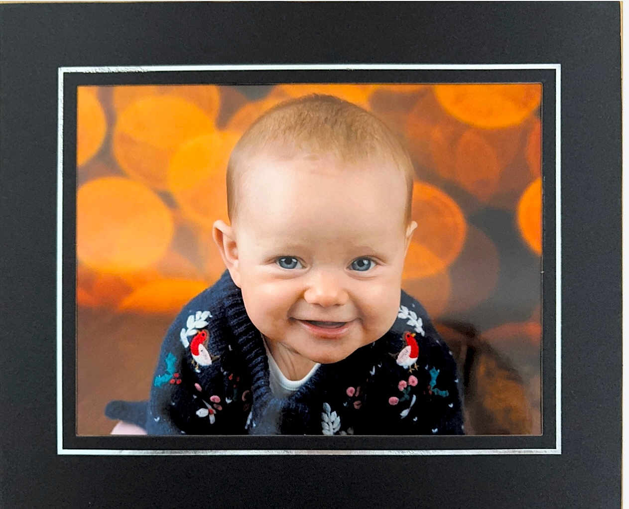 Amelia joined the competition — help win amazing prizes! baby, blue_eyes, smile, sweater, bird_pattern, floral_pattern, portrait, indoor, bokeh, orange_background, close_up, child, cute, happy, framed_photo, cozy_clothing, infant, face, head, person