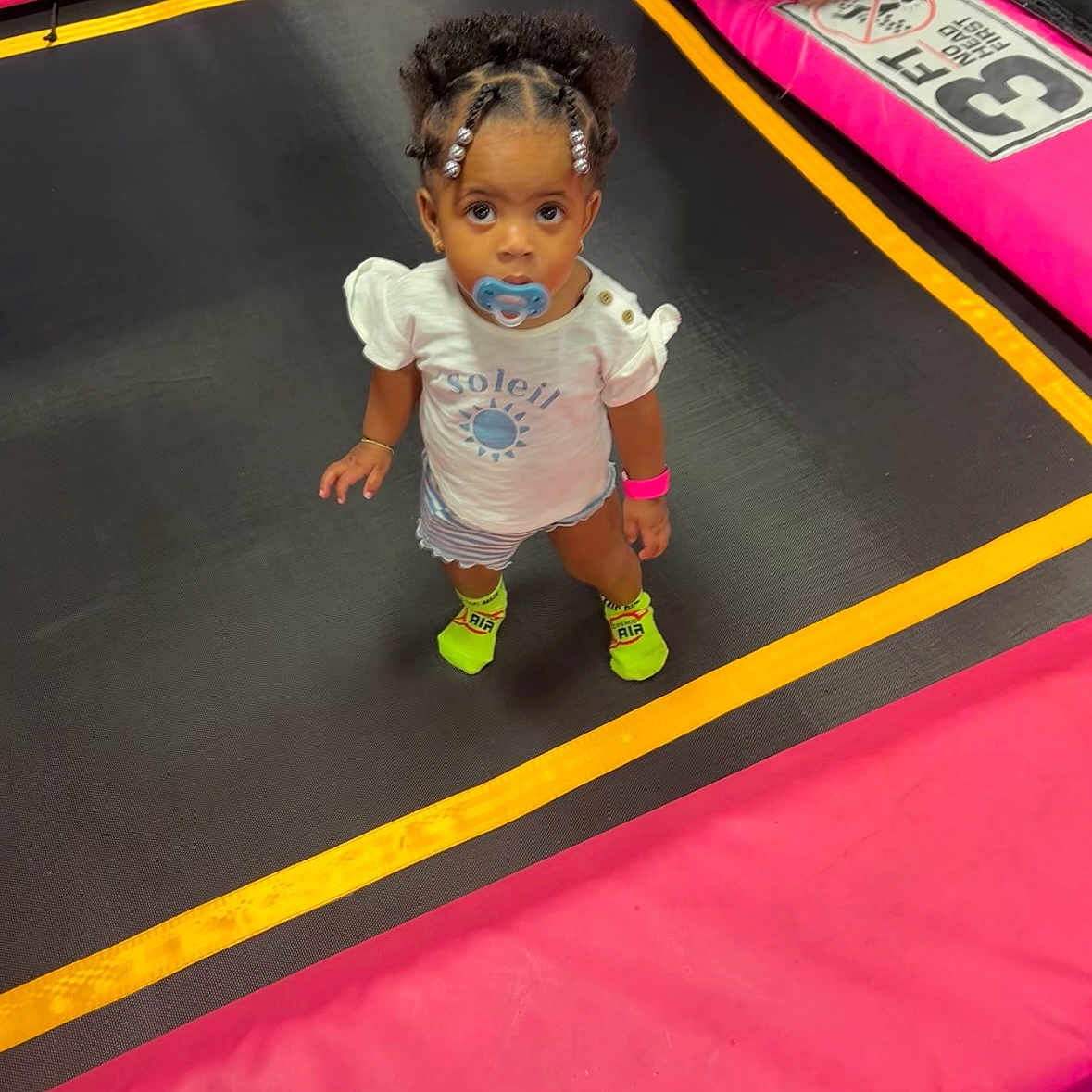 Anilah is registered to the contest to win money with this photo: bracelet, child, curly_hair, cute, footwear, indoor, looking_up, neon_socks, pacifier, pink, play_area, shorts, small_child, standing, toddler, trampoline, vibrant_colors, white_shirt, yellow, young