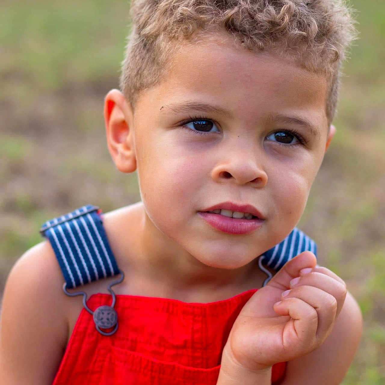 Elijah joined the competition — help win amazing prizes! background_blur, blue_straps, boy, casual_clothing, child, close_up, curly_hair, expression, eyes, face, grass, hand, natural_light, outdoor, portrait, red_overalls, skin, summer, thoughtful, young
