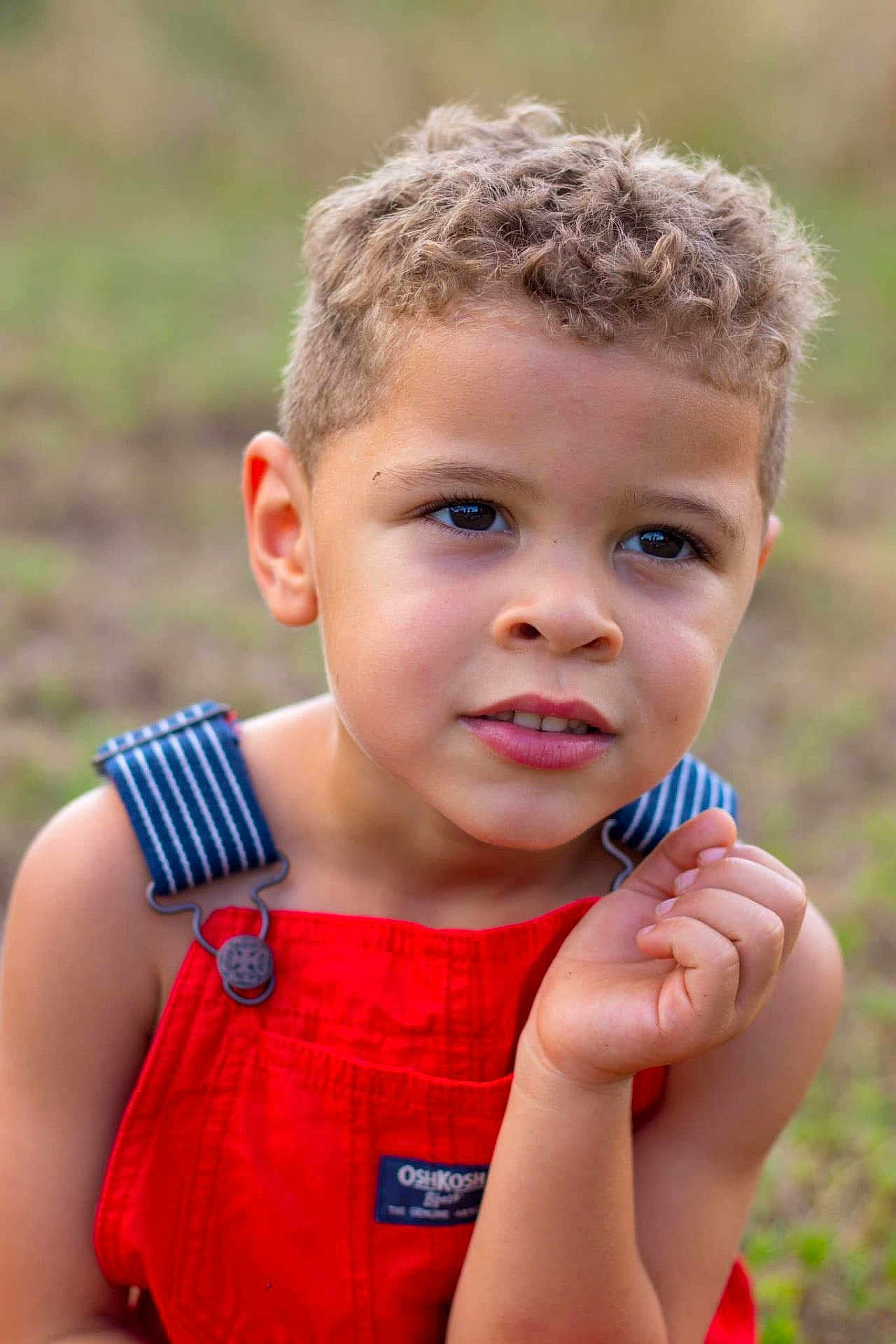 Elijah joined the competition — help win amazing prizes! child, curly_hair, red_overalls, blue_straps, outdoor, portrait, close_up, thoughtful, young, boy, natural_light, grass, summer, skin, face, eyes, hand, casual_clothing, expression, background_blur