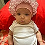 baby, infant, child, pink_bonnet, white_onesie, red_background, cute, chubby_cheeks, indoor, portrait, person, young_child, sitting, headwear, clothing, face, skin, arms, expression, curious