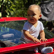 Thaïs a rejoint le concours — aidez-le/la à gagner de superbes lots ! child, toddler, baby, red_car, car_trunk, outdoor, sunlight, shadow, hands, socks, white_shirt, portrait, candid, greenery, bokeh, face, eyes, smile, vehicle, rear_window