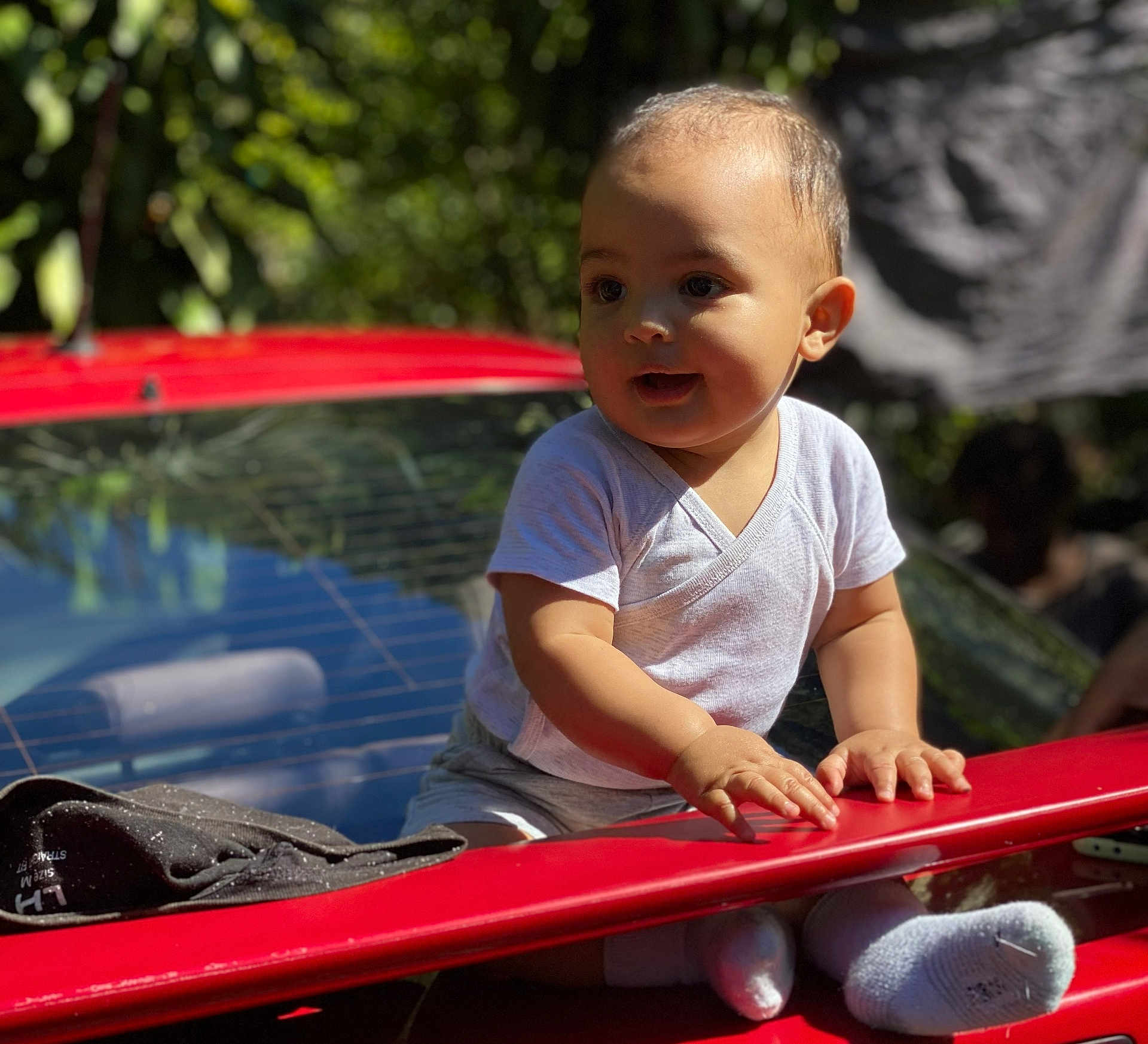 Thaïs a rejoint le concours — aidez-le/la à gagner de superbes lots ! child, toddler, baby, red_car, car_trunk, outdoor, sunlight, shadow, hands, socks, white_shirt, portrait, candid, greenery, bokeh, face, eyes, smile, vehicle, rear_window