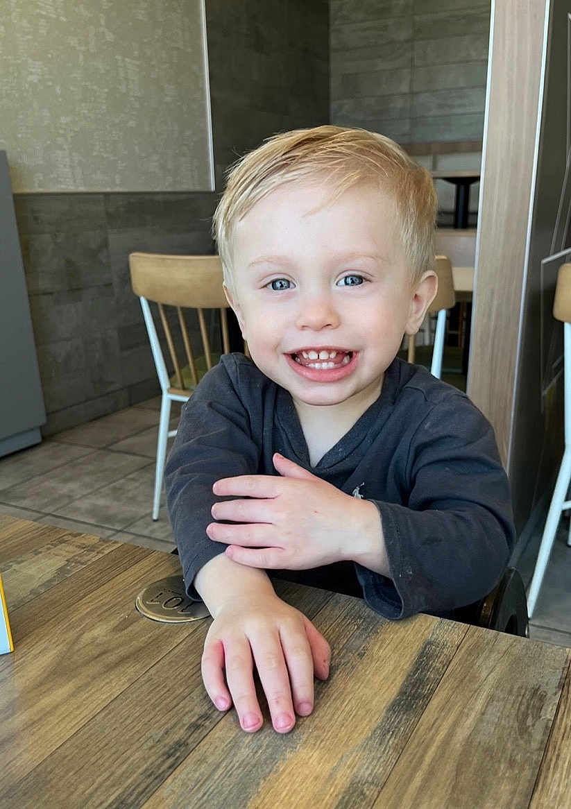 Kyler joined the competition — help win amazing prizes! child, toddler, smiling, blond_hair, blue_eyes, indoor, table, wood, chair, casual_clothing, happy, person, face, hand, cafe, background, furniture, light, portrait, cute