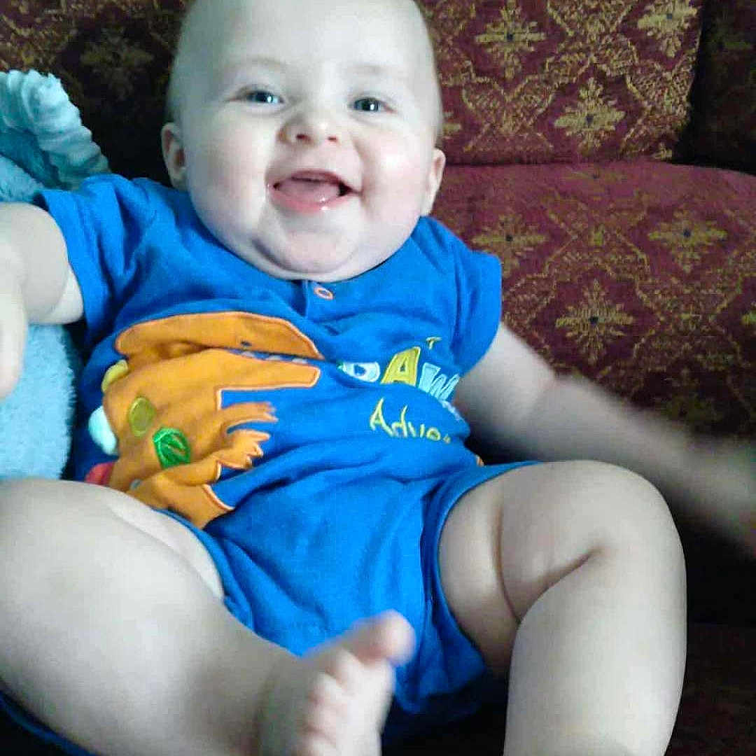 Micah is registered to the contest to win money with this photo: baby, blue_clothing, child, comfort, couch, cute, face, feet, happy, home, indoor, infant, legs, patterned_fabric, person, portrait, relaxed, seated, skin, smiling