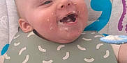 Micah is registered to the contest to win money with this photo: baby, child, high_chair, bib, food, messy_face, spoon, smiling, happy, indoor, feeding, cute, infant, face, head, clothing, pattern, background, toy, seat