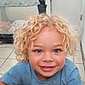 baby_toy, blonde_hair, blue_eyes, casual_clothing, child, childhood, curly_blonde, curly_hair, cute, floor, happy, home, indoor, person, playpen, portrait, smiling, tile, toy, young