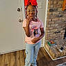 child, girl, glasses, red_bow, beads, peace_sign, pink_shirt, graphic_tee, cargo_pants, white_sneakers, indoor, wood_floor, brick_fireplace, books, electronic_charger, door, pouting, standing, playful, home