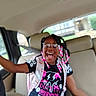 child, smiling, glasses, beads, car_interior, seatbelt, backseat, black_hair, tshirt, pink, joyful, happy, person, hairstyle, casual_clothing, seat, window, daylight, portrait, sitting