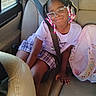 child, girl, car, seatbelt, glasses, braids, beads, purple_shirt, plaid_shorts, backseat, pink_backpack, cherry_pattern, window, sunlight, smile, seated, interior, casual_clothing, happy, portrait