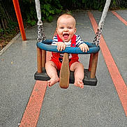 Eden participe au concours pour gagner de l'argent avec cette photo : baby, child, swing, playground, smiling, happy, red_overalls, striped_shirt, outdoor, fun, joy, greenery, pavement, play, summer, park, hands, feet, blue_swing, metal_chains
