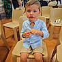 child, boy, blue_shirt, shorts, plastic_chair, indoor, people, group, gathering, event, floor, beige_chair, holding, orange_object, casual_clothing, short_hair, seated, looking_at_camera, mixed_ethnicity, daylight