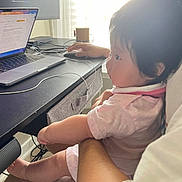 Isla is registered to the contest to win money with this photo: baby, child, infant, person, adult, laptop, computer_mouse, desk, window, curtains, technology, home_office, cable, screen, arm, leg, pink_clothing, indoor, light, working