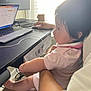 baby, child, infant, person, adult, laptop, computer_mouse, desk, window, curtains, technology, home_office, cable, screen, arm, leg, pink_clothing, indoor, light, working