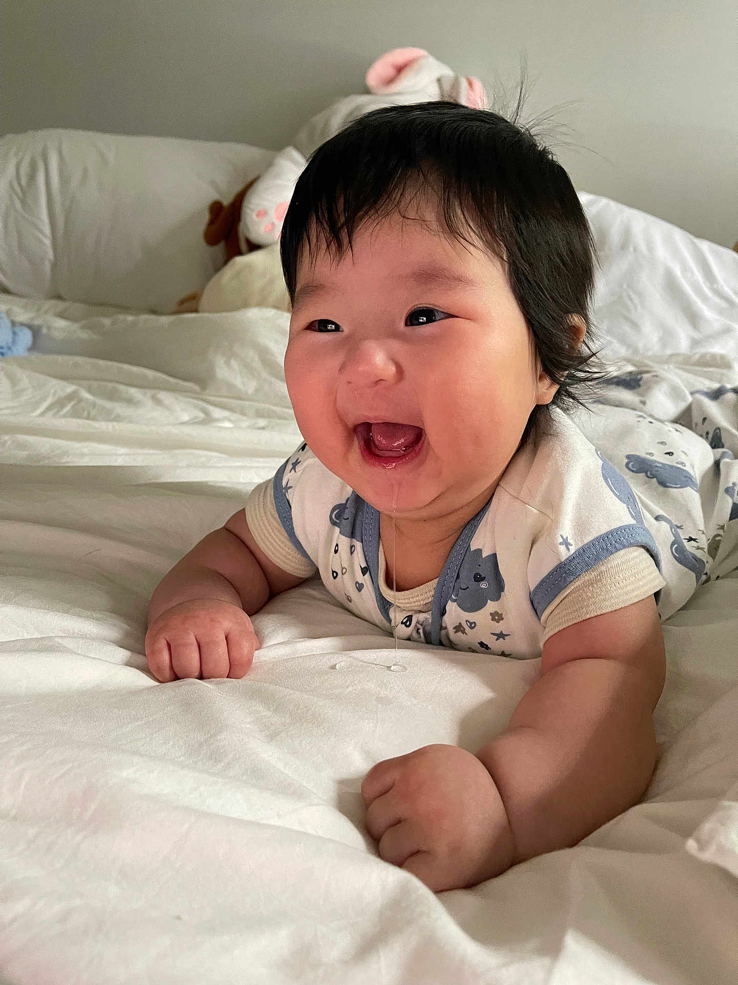 Isla is registered to the contest to win money with this photo: baby, infant, smiling, drooling, bed, bedding, plush_toy, white, blue, clothing, happy, cute, child, indoors, person, portrait, lying_down, soft, home, cozy