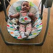 Athena is registered to the contest to win money with this photo: baby, child, swing, plush_toy, cow_toy, pink_clothing, socks, floor, wood_floor, baby_seat, smiling, indoor, infant, person, cute, comfort, seatbelt, colorful, happy, toy