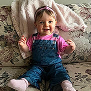 Athena joined the competition — help win amazing prizes! baby, blanket, child, clothing, couch, cozy, cute, denim_overalls, floral_pattern, furniture, happy, headband, indoor, infant, person, pink_shirt, portrait, seated, smiling, socks