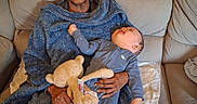William is registered to the contest to win money with this photo: elderly_person, baby, blanket, plush_toy, couch, living_room, cozy, clothing, person, indoor, family, comfort, smile, holding, seated, relaxation, home, infant, soft_lighting, tender_moment
