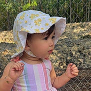 Cleidys is registered to the contest to win money with this photo: baby, chain_link_fence, child, curious, face, fence, floral_hat, garden, hands, nature, outdoor, pink_clothing, portrait, profile, standing, striped_clothing, sunhat, sunlight, toddler, trees