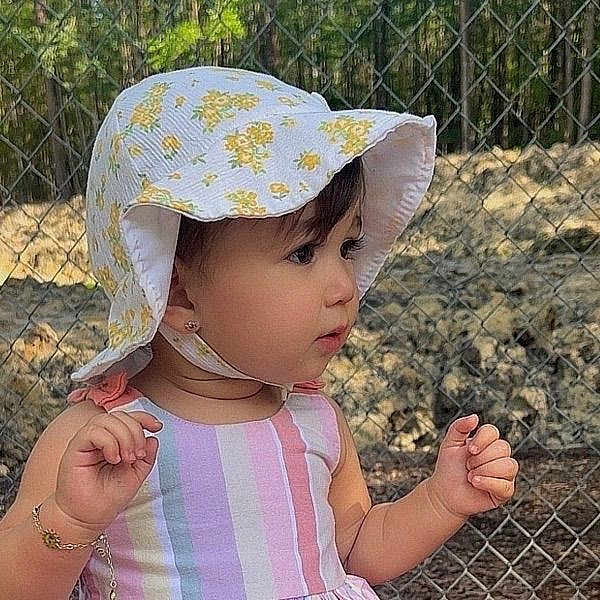 Cleidys is registered to the contest to win money with this photo: baby, chain_link_fence, child, curious, face, fence, floral_hat, garden, hands, nature, outdoor, pink_clothing, portrait, profile, standing, striped_clothing, sunhat, sunlight, toddler, trees