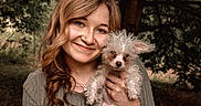 Pebbles joined the competition — help win amazing prizes! woman, person, dog, puppy, pet, portrait, smiling, outdoors, forest, woodland, trees, nature, curly_fur, scruffy_dog, brown_hair, long_hair, gray_sweater, holding_pet, cuddling, natural_light