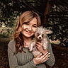 woman, person, dog, puppy, pet, portrait, smiling, outdoors, forest, woodland, trees, nature, curly_fur, scruffy_dog, brown_hair, long_hair, gray_sweater, holding_pet, cuddling, natural_light