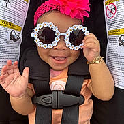 Kailani is registered to the contest to win money with this photo: baby, child, sunglasses, headband, pink_flower, car_seat, smile, happy, waving_hand, bracelet, black_straps, orange_shirt, infant, cute, accessory, portrait, seatbelt, safety, closeup, fashion