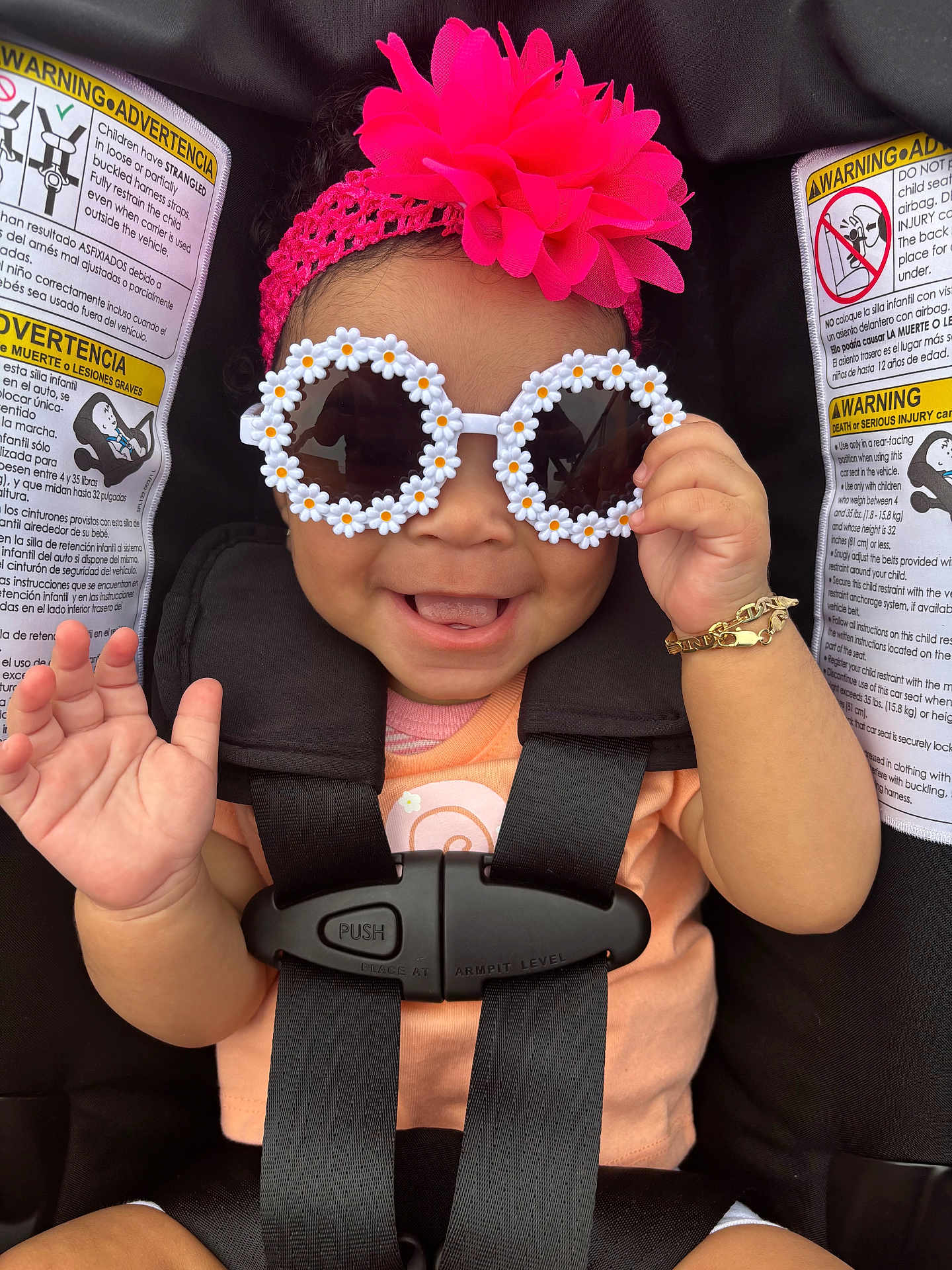Kailani is registered to the contest to win money with this photo: baby, child, sunglasses, headband, pink_flower, car_seat, smile, happy, waving_hand, bracelet, black_straps, orange_shirt, infant, cute, accessory, portrait, seatbelt, safety, closeup, fashion