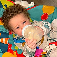Greyson joined the competition — help win amazing prizes! baby, bottle, child, curly_hair, drink, feeding, hand, infant, indoors, play_seat, plush, seat, sitting, toy, wood_floor, young_child, colorful, clothing, pajamas, cute