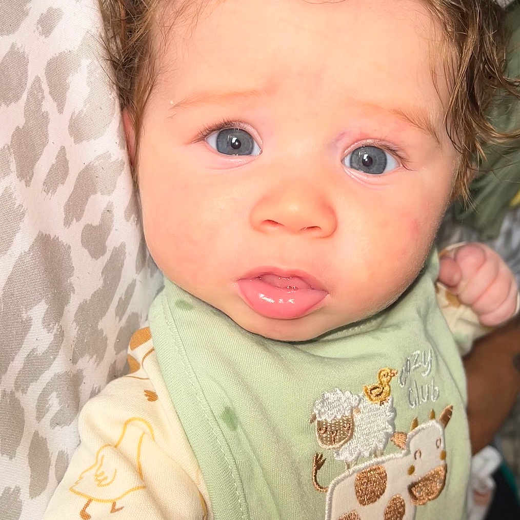 Greyson joined the competition — help win amazing prizes! baby, infant, blue_eyes, green_bib, animal_embroidery, cream_outfit, duck_prints, curly_hair, close_up, face, child, cute, portrait, indoor, patterned_background, clothing, young_child, person, expression, soft_lighting