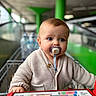 baby, child, pacifier, shopping_cart, sweater, blue_eyes, indoor, person, cute, infant, portrait, hand, blurred_background, retail, store, shopping, seat, clothing, young_child, face