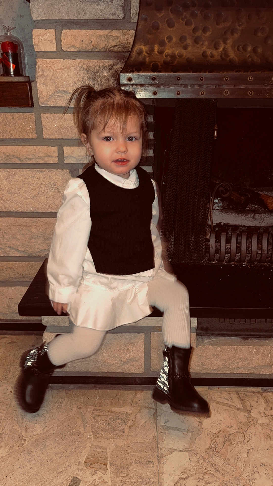 Inaya participe au concours pour gagner de l'argent avec cette photo : bench, black_vest, boots, brick_wall, child, clothing, cozy, dress, face, fireplace, floor_tiles, girl, hearth, indoor, pigtails, portrait, sitting, smiling, toddler, white_tights
