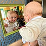 baby, mirror, reflection, smile, indoor, face, infant, clothing, bib, hand, person, wallpaper, pattern, frame, adult, furniture, light, curiosity, portrait, holding