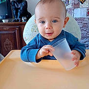Samson participe au concours pour gagner de l'argent avec cette photo : baby, child, blue_sweater, high_chair, plastic_cup, indoor, furniture, gorilla_figurine, shelf, cabinet, face, person, smiling, cute, home, decor, light, portrait, hand, table
