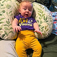 Myles is registered to the contest to win money with this photo: baby, infant, child, pillow, leaf_pattern, purple_shirt, yellow_pants, viking_logo, clothing, cute, expression, indoors, fabric, blanket, comfort, sitting, face, human, smiling, soft