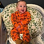 baby, child, infant, orange_clothing, pumpkin_pattern, boots, baby_seat, cushion, leaf_pattern, indoor, happy, smiling, face, hands, feet, person, cute, comfortable, surprised, expression