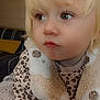 toddler, child, blonde_hair, curious_eyes, leopard_print, fuzzy_vest, bear_design, cozy_clothing, indoor, couch, striped_pillow, yellow_cushion, closeup, portrait, soft_texture, casual_wear, young_child, sitting, face, person