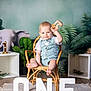 baby, child, wicker_chair, wooden_toy, elephant, green_leaves, milestone, one, letters, smiling, cute, indoor, portrait, floor_rug, clothing, shorts, vest, celebration, happy, toddler