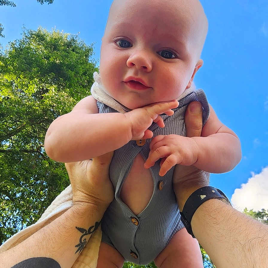 Grayson is registered to the contest to win money with this photo: arms, baby, blue_sky, child, cute, greenery, happy, holding, infant, nature, outdoor, person, playing, portrait, sky, summer, sunlight, tattoo, trees, watch