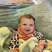 Makenna joined the competition — help win amazing prizes! baby, smiling, blanket, curly_hair, couch, cushion, infant, happy, child, indoors, person, cute, face, clothing, patterned_blanket, soft_texture, playful, young, portrait, seated