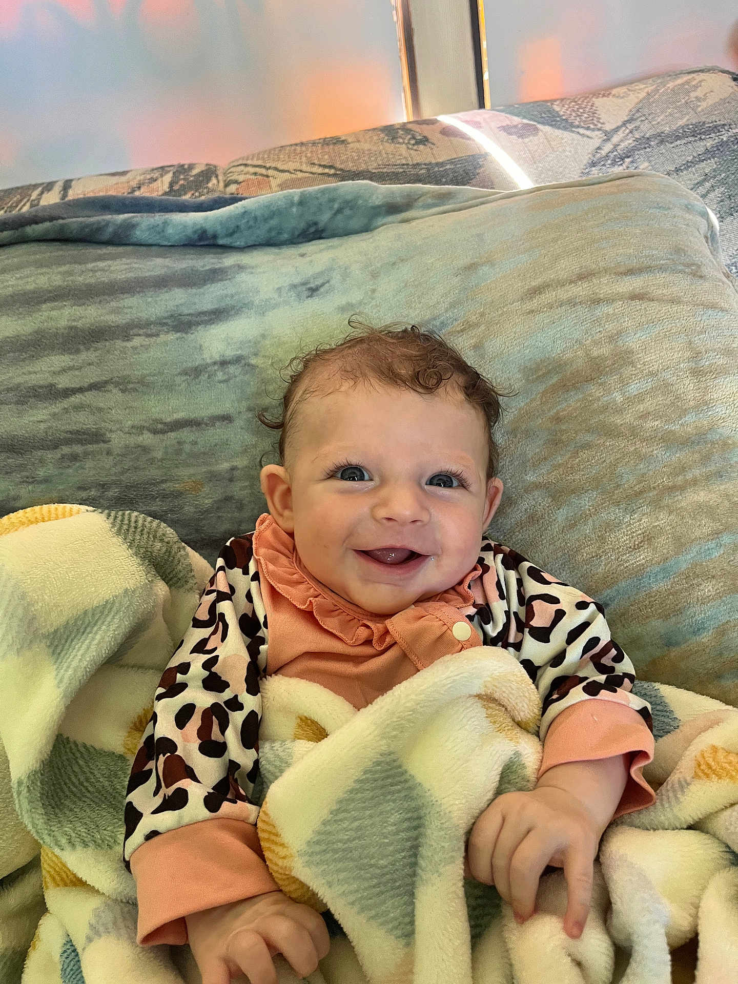 Makenna joined the competition — help win amazing prizes! baby, smiling, blanket, curly_hair, couch, cushion, infant, happy, child, indoors, person, cute, face, clothing, patterned_blanket, soft_texture, playful, young, portrait, seated