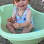 bathtub, blue_eyes, child, daylight, hair_clip, happy, messy_face, necklace, outdoor, pebbled_ground, person, plastic_tub, playtime, sitting, smiling, summer, swimsuit, toddler, toy_ball, water